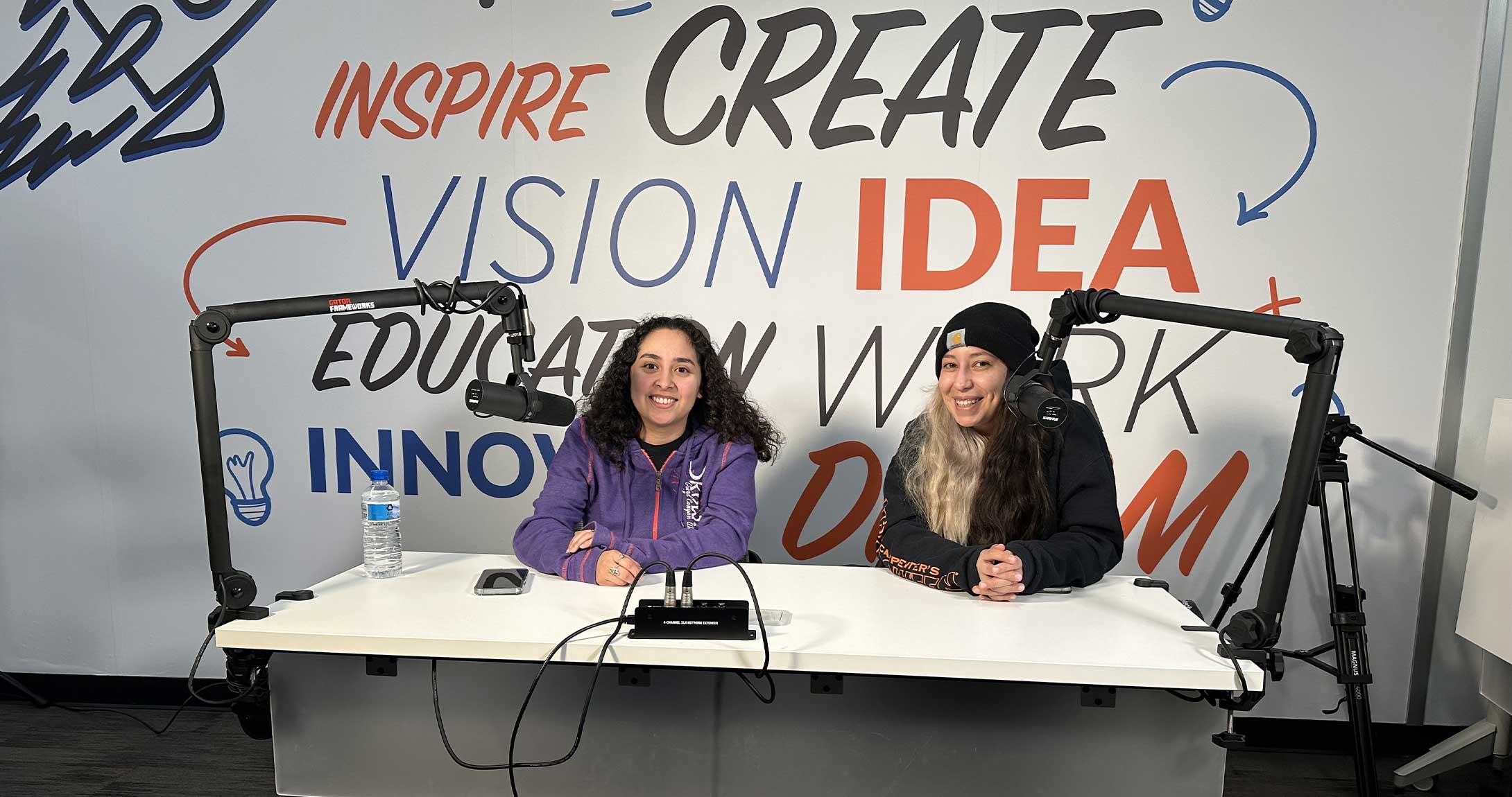two students smile while sitting at the podcast table 