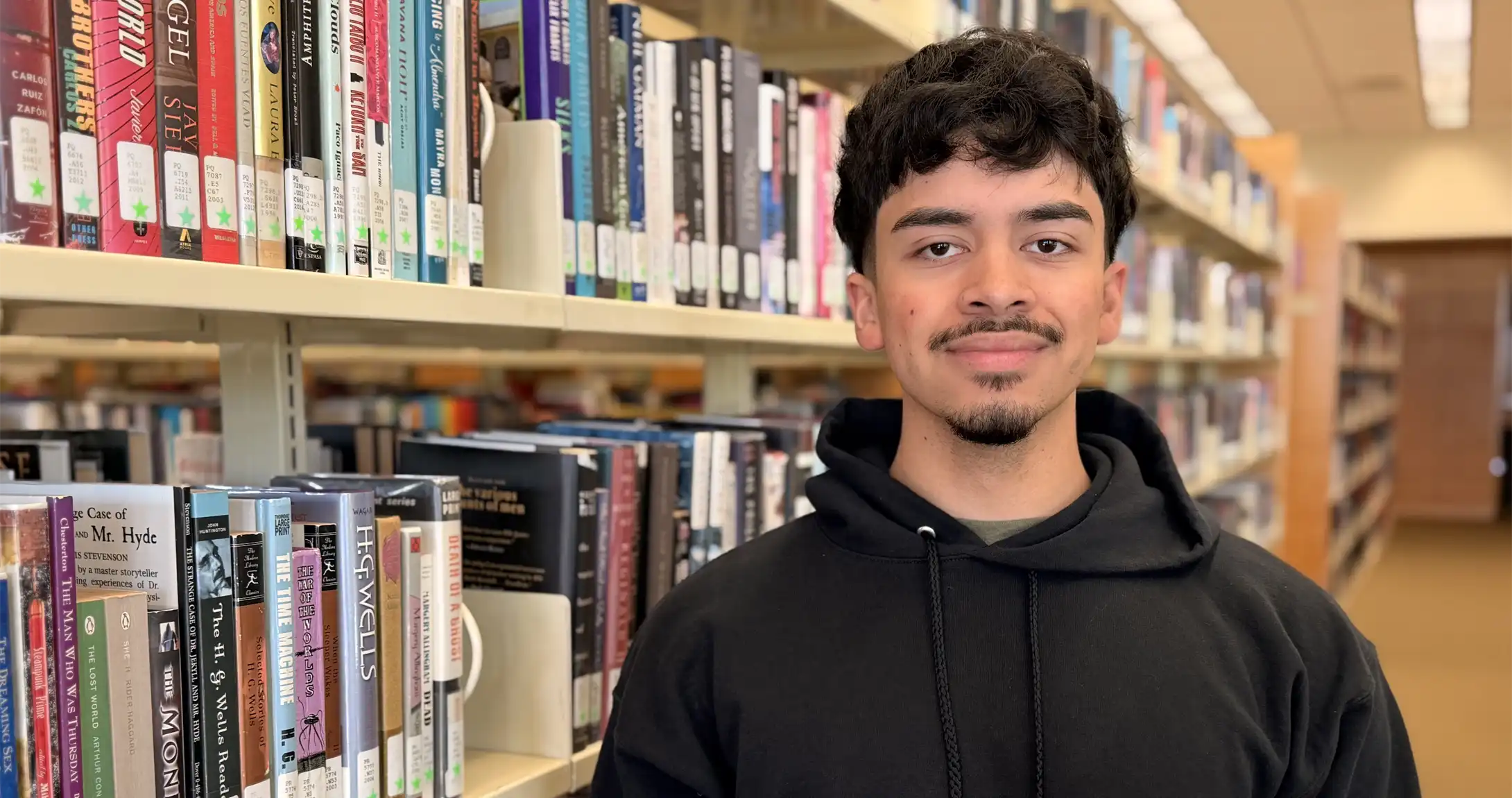 Cesar Vasquez standing among books in the UTPB library. 